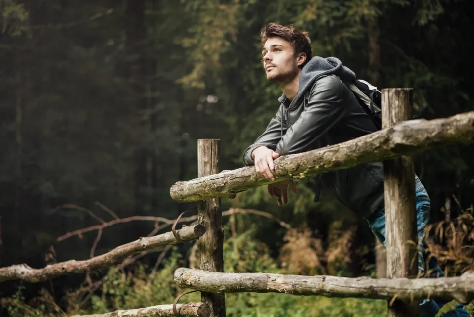 A young man in a forest leaning on a fence look out.